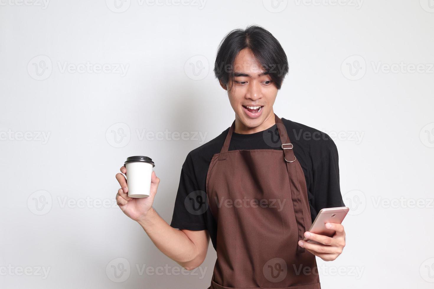 Portrait of attractive Asian barista man in brown apron holding disposable paper coffee cup while using mobile phone. Isolated image on white background photo