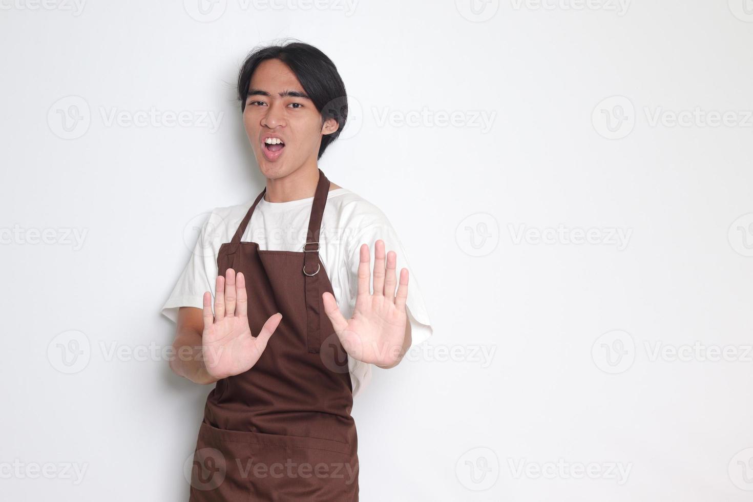Portrait of attractive Asian barista man in brown apron forming a hand gesture to avoid something. Isolated image on white background photo