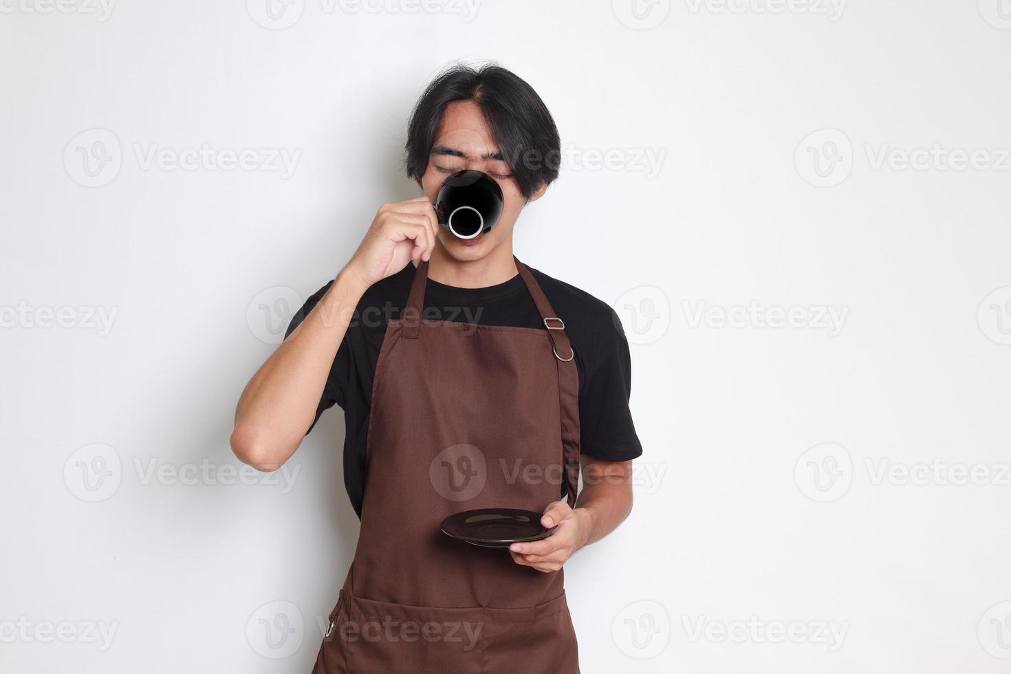Portrait of attractive Asian barista man in brown apron drinking a cup of coffee while holding the saucer. Isolated image on white background photo