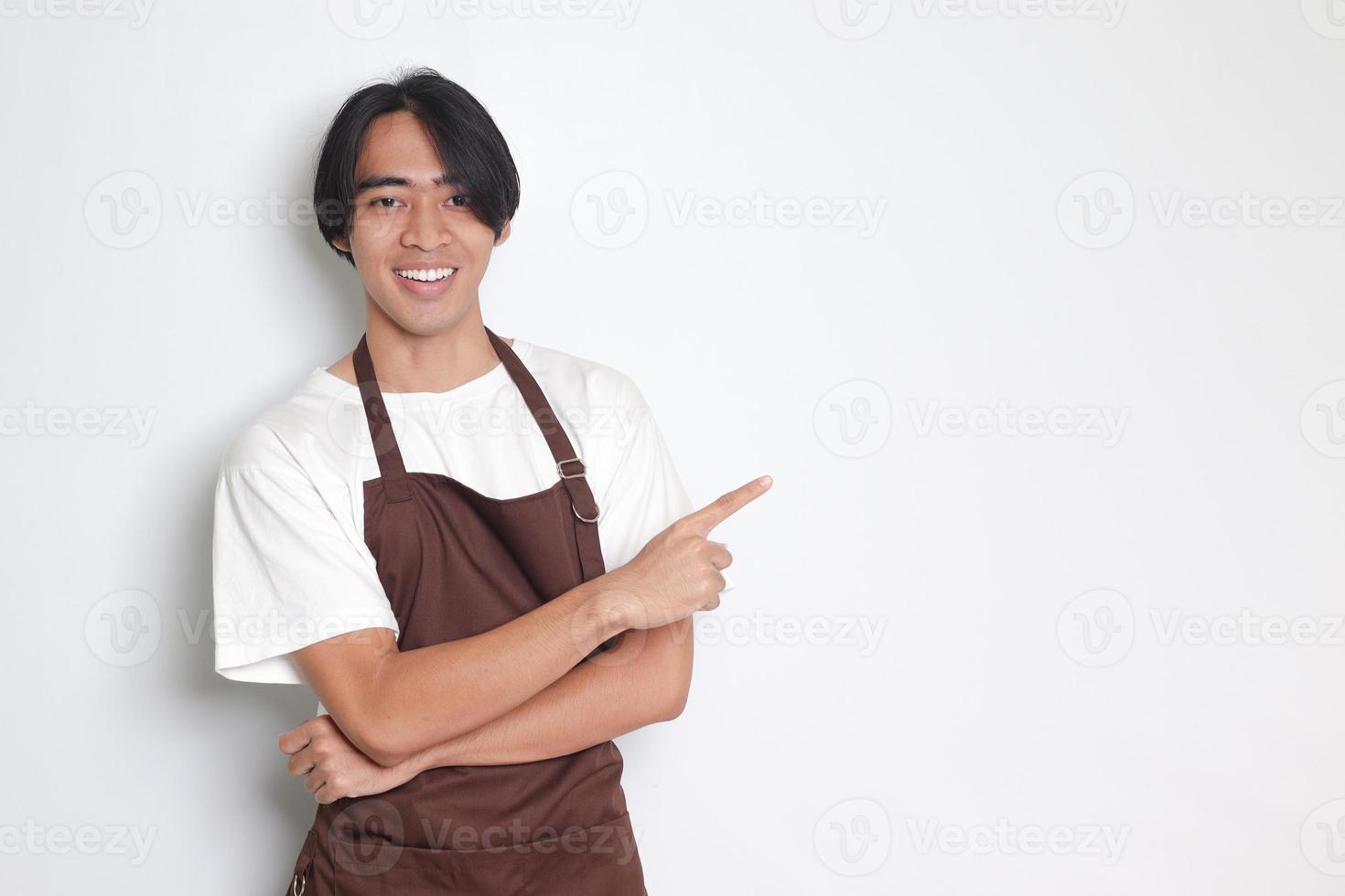 Portrait of attractive Asian barista man in brown apron showing product, pointing at something with hands. Advertising concept. Isolated image on white background photo