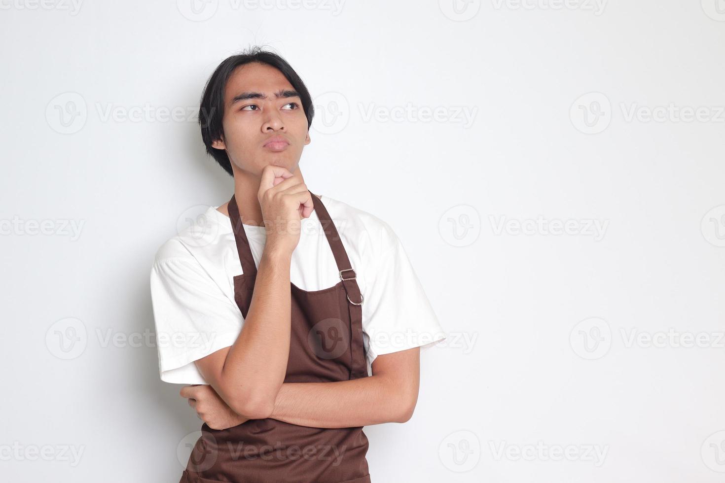 Portrait of attractive Asian barista man in brown apron thinking about question with hand on chin. Isolated image on white background photo