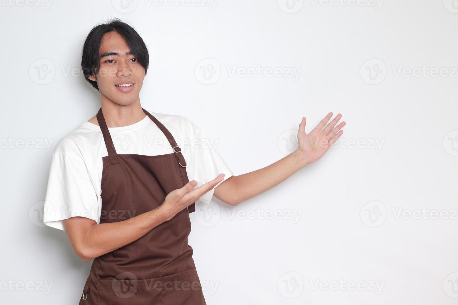 Portrait of attractive Asian barista man in brown apron showing product, pointing at something with hands. Advertising concept. Isolated image on white background photo