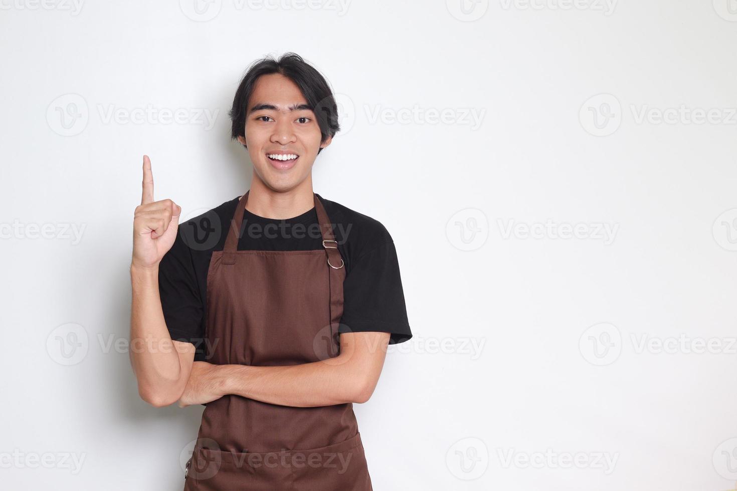 Portrait of attractive Asian barista man in brown apron showing product, pointing at something with hands. Advertising concept. Isolated image on white background photo