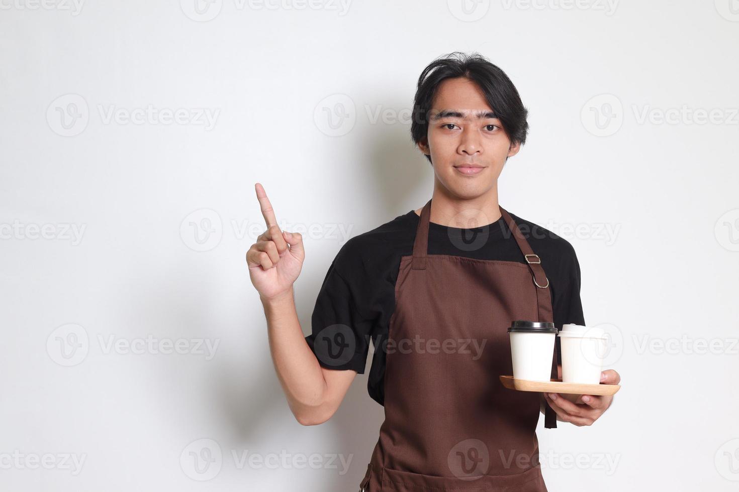 Portrait of attractive Asian barista man in brown apron holding take away paper coffee cup with wooden tray to serve while pointing to the side. Isolated image on white background photo