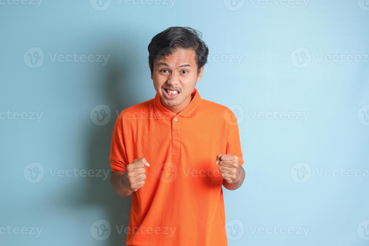 annoyed Asian man in orange shirt making angry hand gesture with fingers. Isolated image on blue background photo