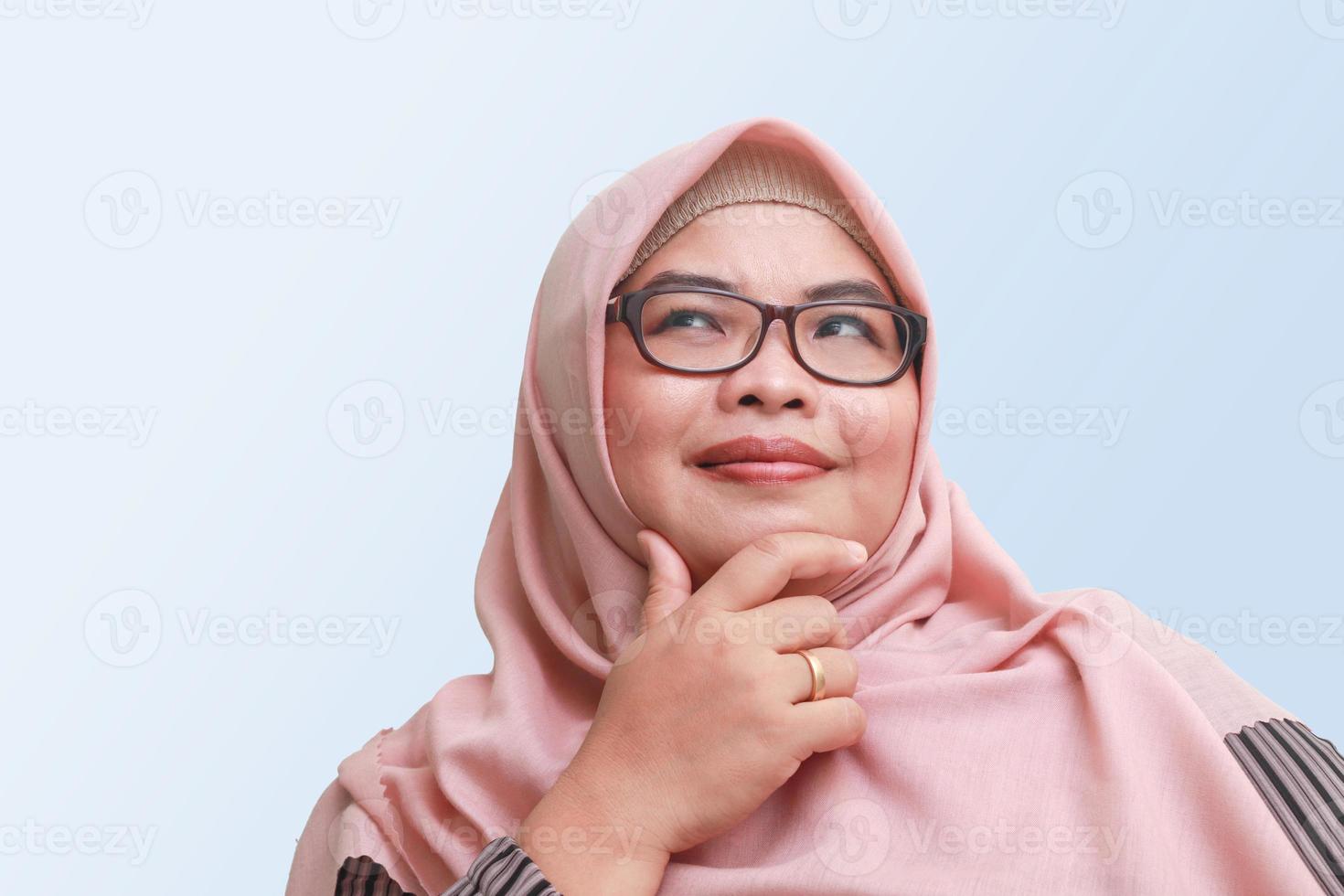 retrato de alegre asiático mujer con hiyab, en pie en contra azul fondo, pensando acerca de pregunta con mano en barbilla foto