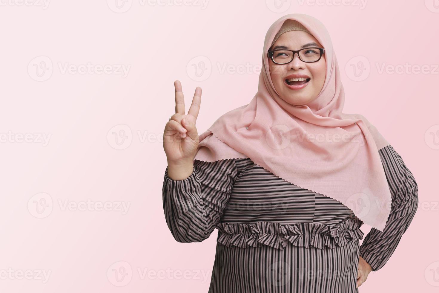 Portrait of cheerful Asian woman with hijab, making fun face with peace sign hand gesture. Advertising concept. Isolated image on pink background photo