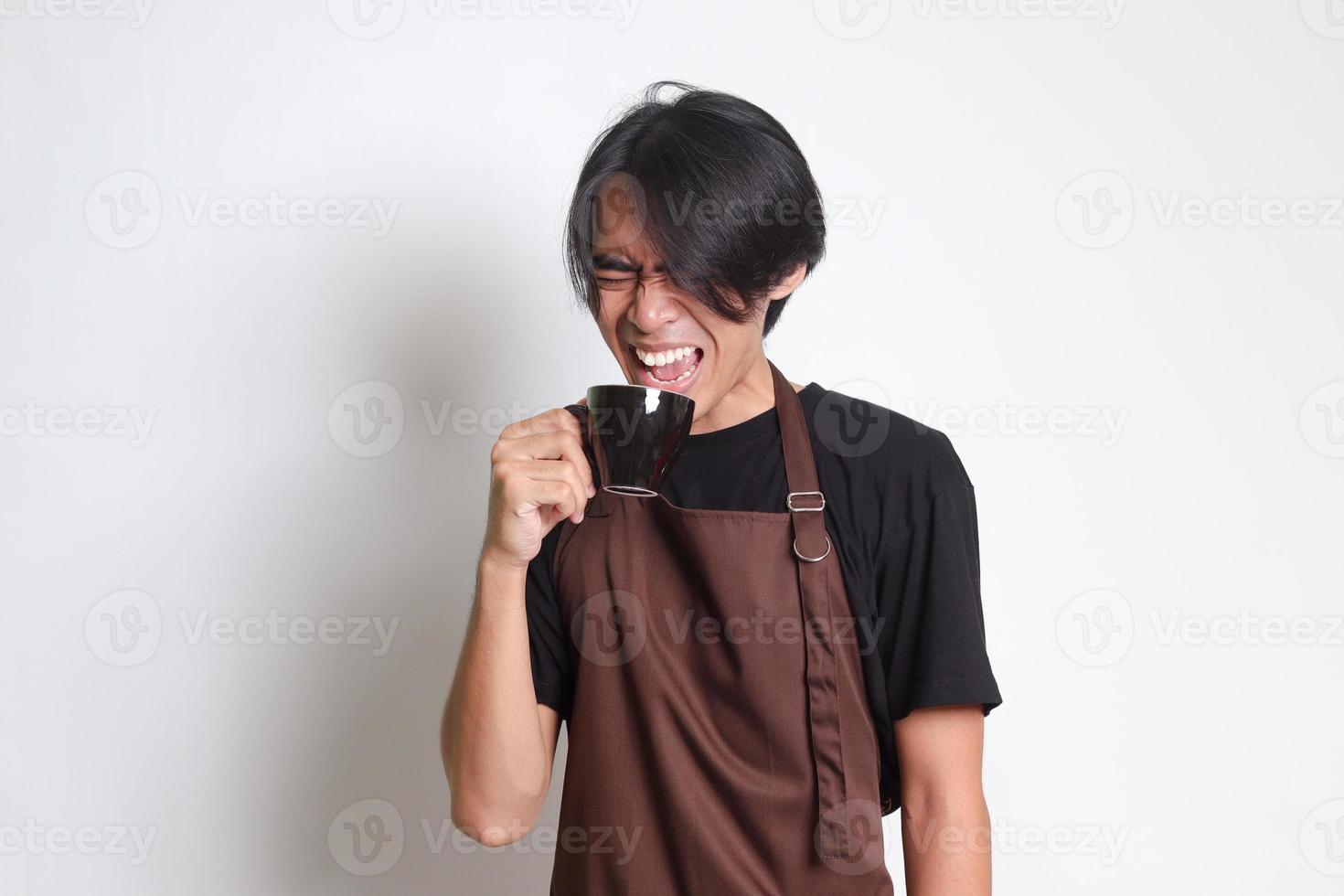 retrato de atractivo asiático barista hombre en marrón delantal haciendo desagradable cara mientras Bebiendo un taza de café. aislado imagen en blanco antecedentes foto