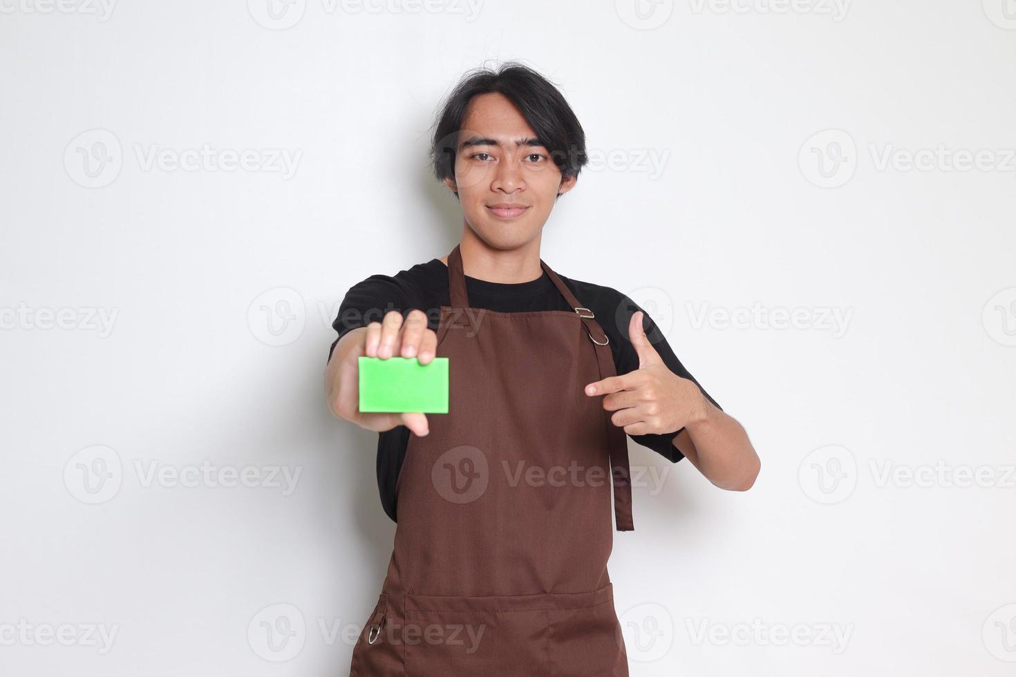 retrato de atractivo asiático barista hombre en marrón delantal demostración modelo de crédito tarjeta y señalando en él. aislado imagen en blanco antecedentes foto