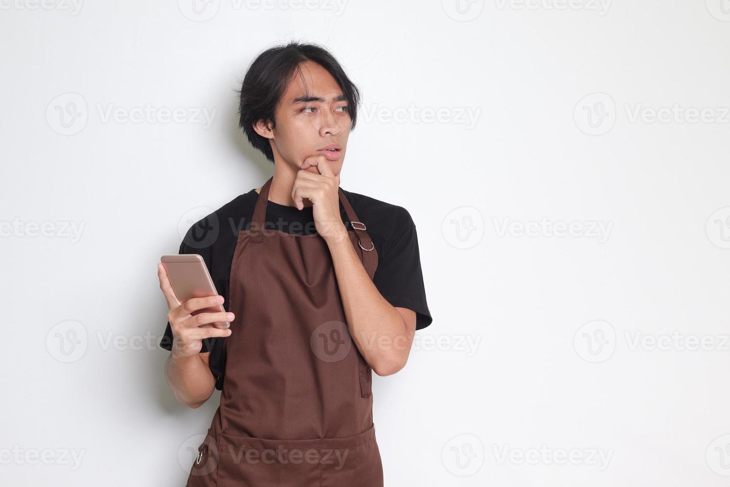 Portrait of attractive Asian barista man in brown apron thinking about question and answer with hand on chin while holding smartphone. Isolated image on white background photo