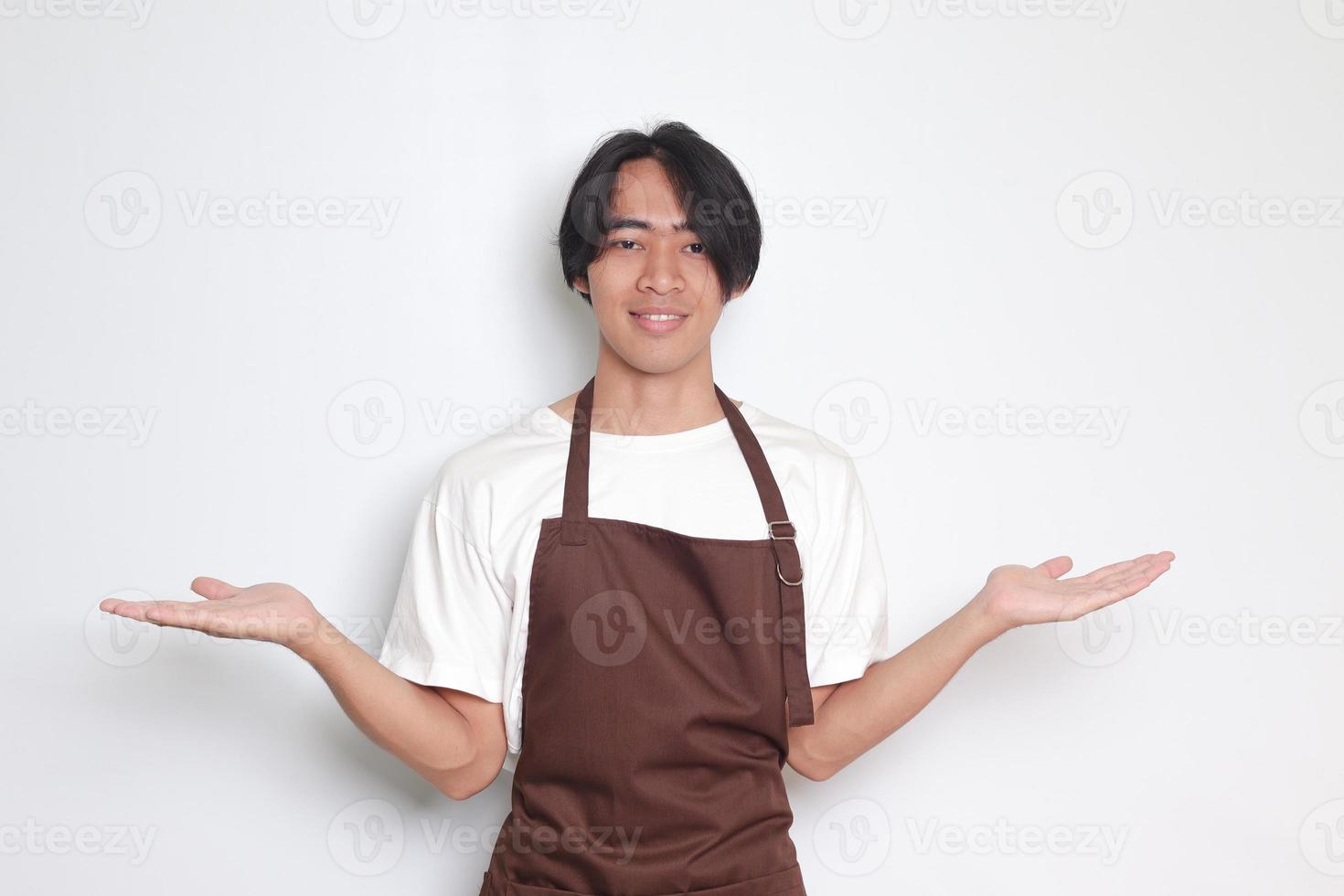 Portrait of attractive Asian barista man in brown apron choosing between 2 different options, holding two hands with empty space. Decision and choice concept. Isolated image on white background photo