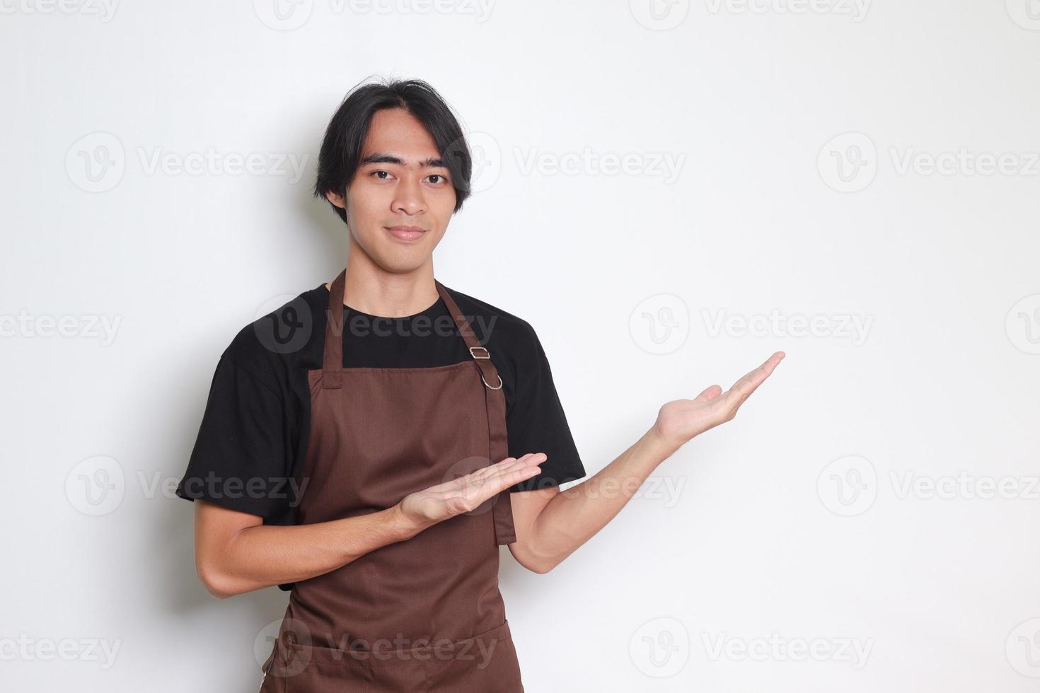 retrato de atractivo asiático barista hombre en marrón delantal demostración producto, señalando a alguna cosa con manos. publicidad concepto. aislado imagen en blanco antecedentes foto