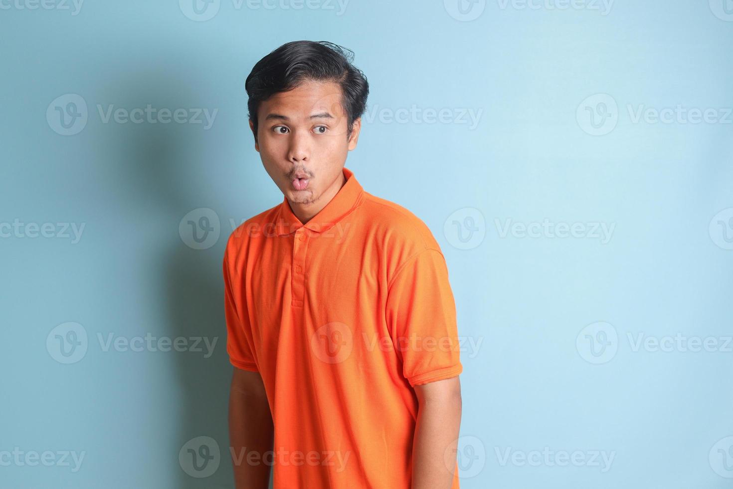 surprised Asian man in orange shirt standing against blue background, showing shocked expression and looking aside photo