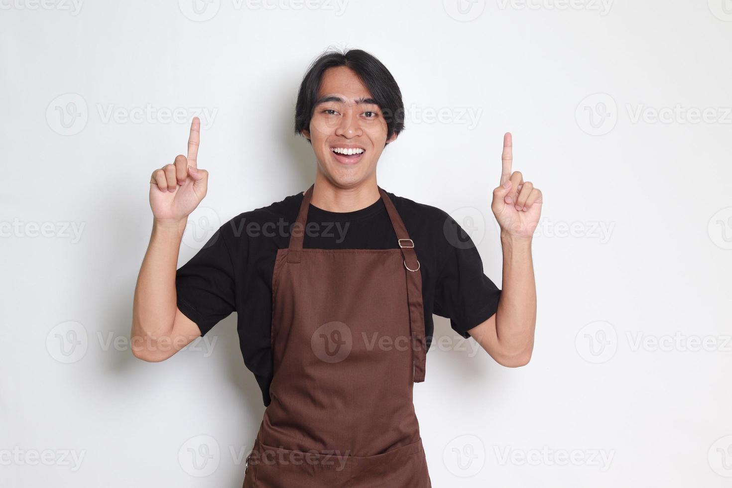 Portrait of attractive Asian barista man in brown apron showing product, pointing at something with hands. Advertising concept. Isolated image on white background photo