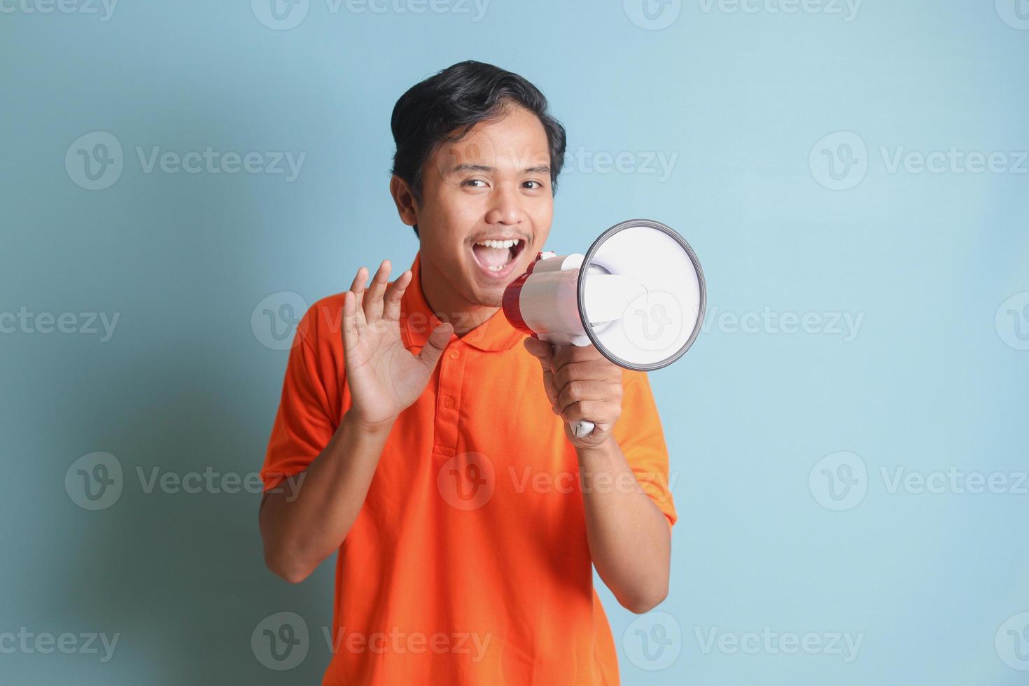 Portrait of attractive Asian man in orange shirt speaking louder using megaphone, promoting product. Advertising concept. Isolated image on blue background photo