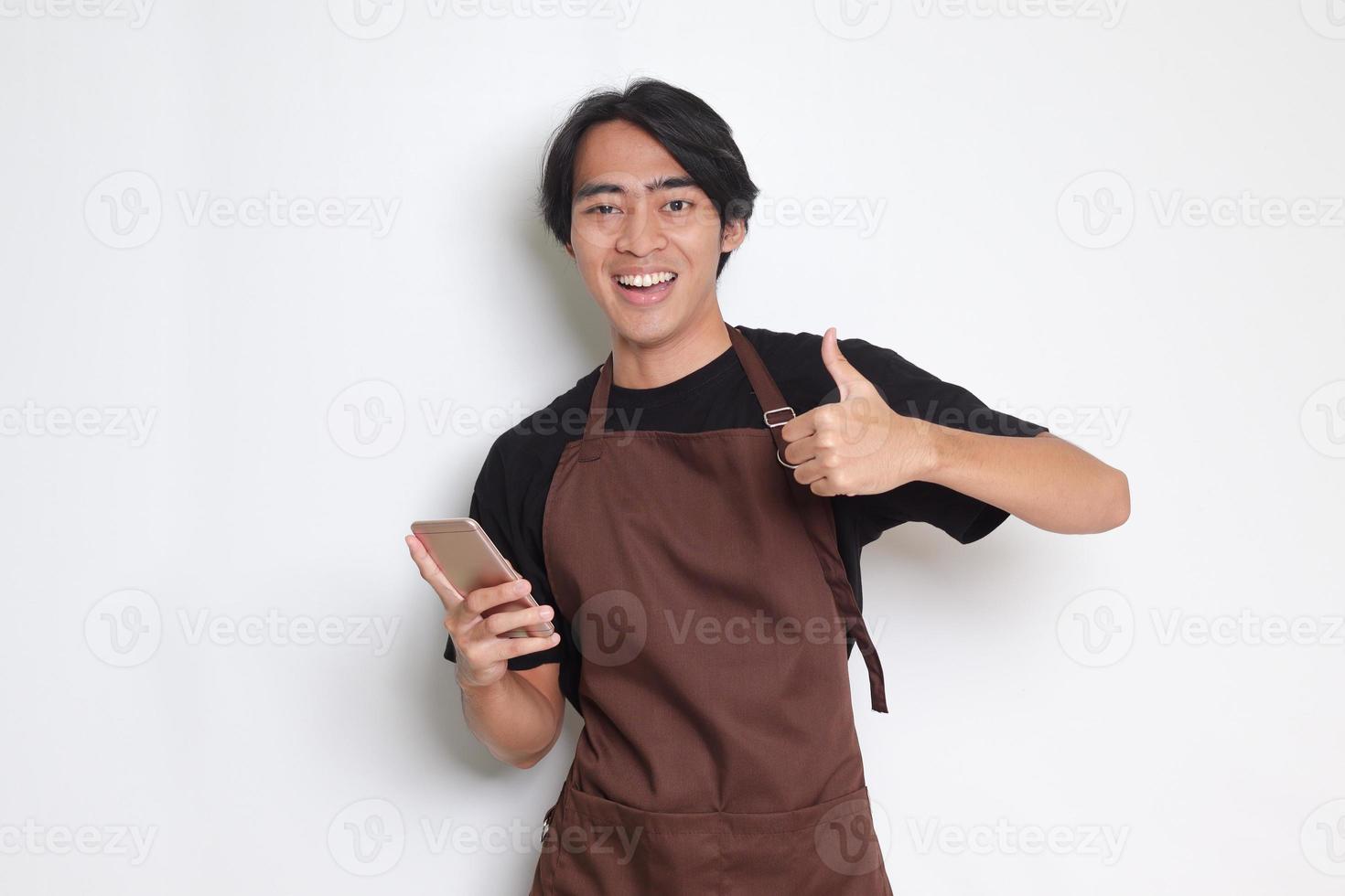 Portrait of attractive Asian barista man in brown apron showing thumb up hand gesture while holding a mobile phone. Advertising concept. Isolated image on white background photo