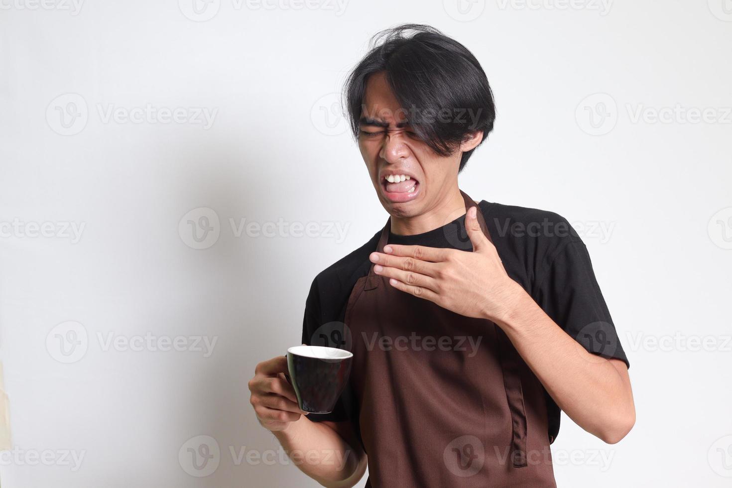 Portrait of attractive Asian barista man in brown apron making unpleasant face while drinking a cup of coffee. Isolated image on white background photo