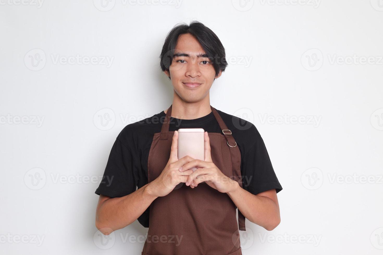 Portrait of attractive Asian barista man in brown apron holding mobile phone while looking at camera. Isolated image on white background photo
