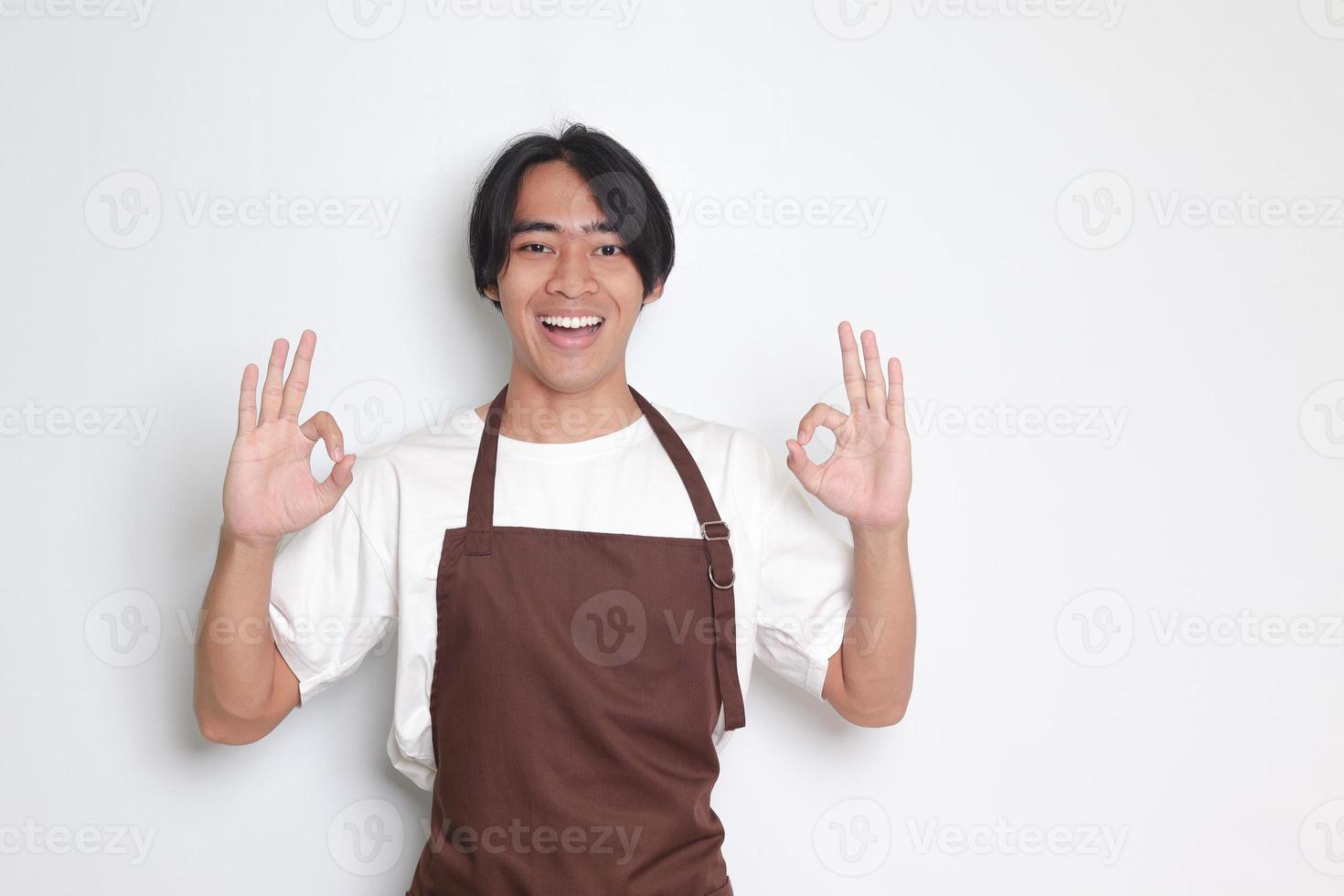 Portrait of attractive Asian barista man in brown apron showing ok hand gesture and smiling looking at camera. Advertising concept. Isolated image on white background photo