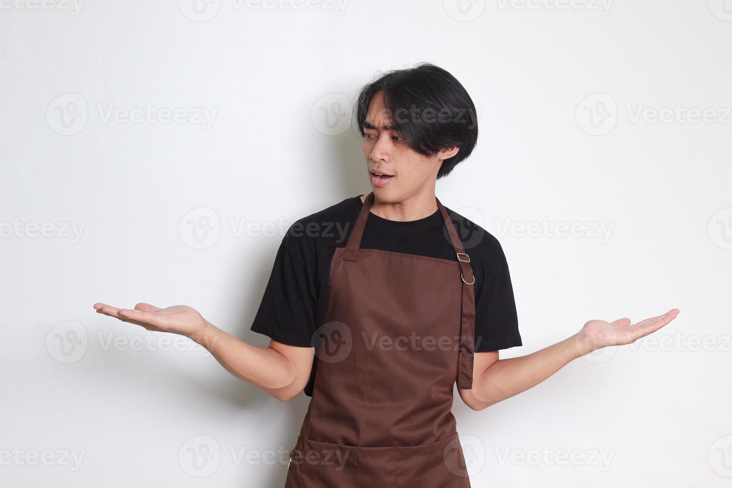 Portrait of attractive Asian barista man in brown apron choosing between 2 different options, holding two hands with empty space. Decision and choice concept. Isolated image on white background photo