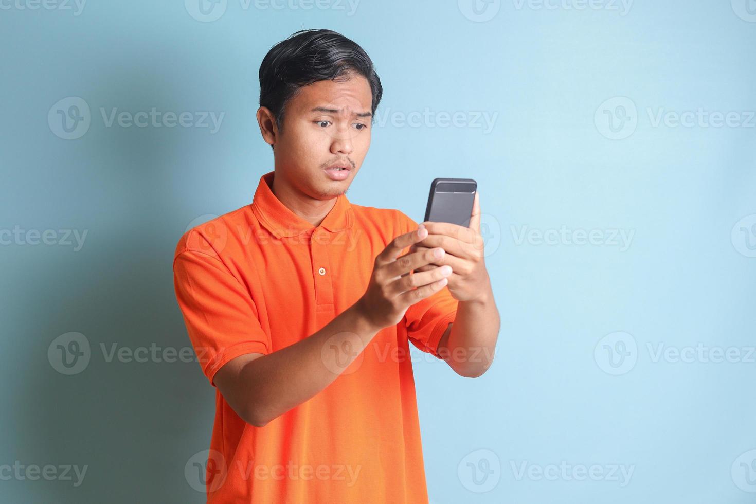 unhappy Asian man in orange shirt holding mobile phone with sad expression on face. Isolated image on blue background photo