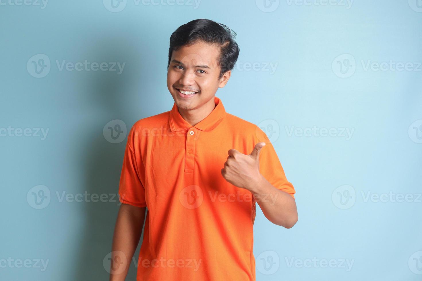 atractivo asiático hombre en naranja camisa con pulgar señalando lejos en vacío espacio. publicidad concepto. aislado imagen en azul antecedentes foto