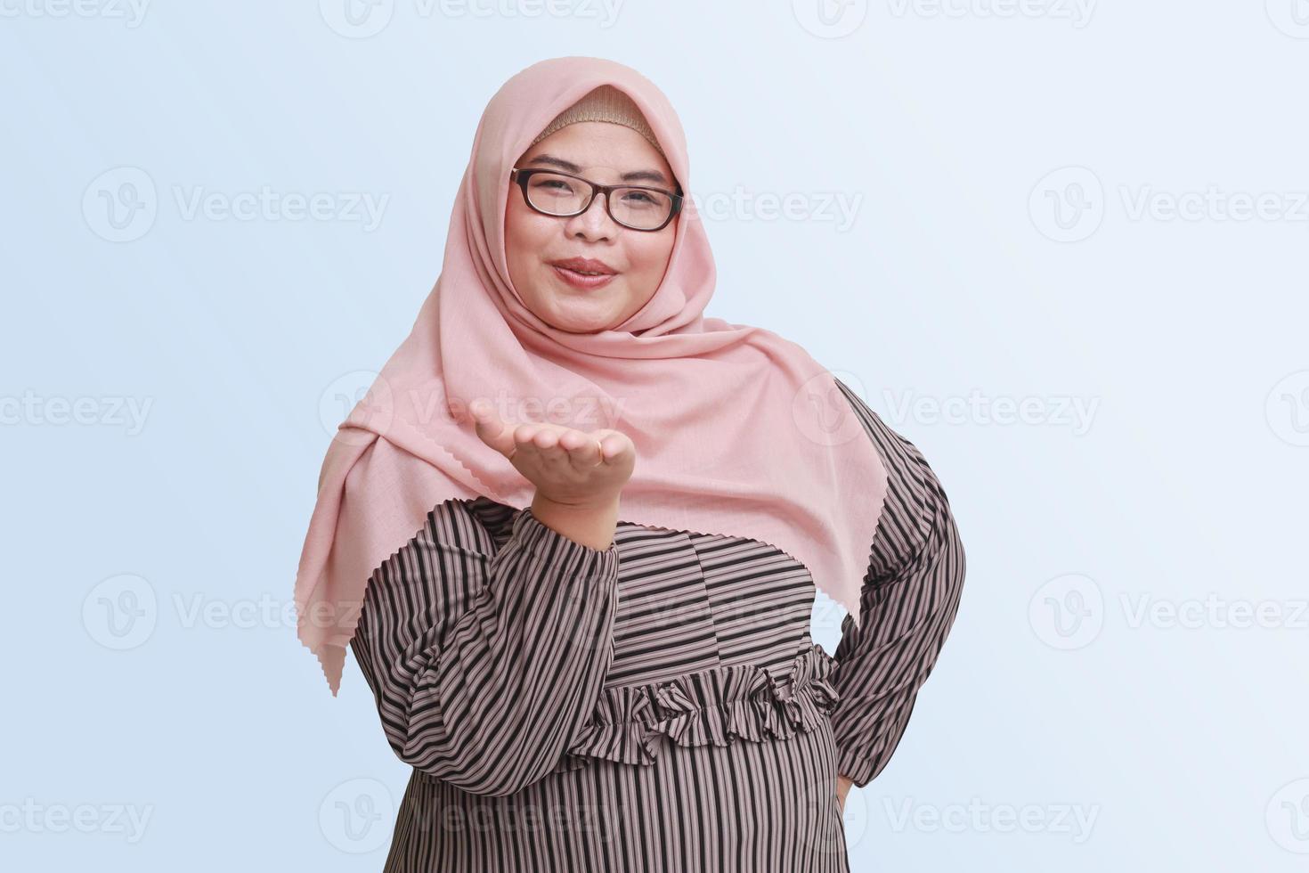 Portrait of cheerful Asian woman with hijab, showing something on empty hand. Advertising concept. Isolated image on pink background photo