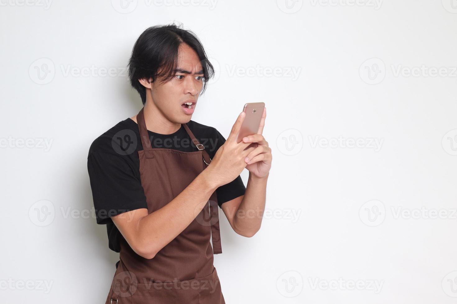 Portrait of shocked Asian barista man in brown apron looking at his mobile phone with surprised expression. Advertising concept. Isolated image on white background photo
