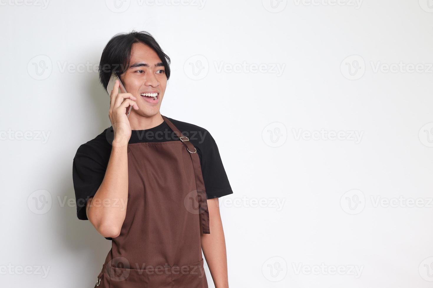 Portrait of attractive Asian barista man in brown apron having conversation while calling someone on mobile phone. Isolated image on white background photo