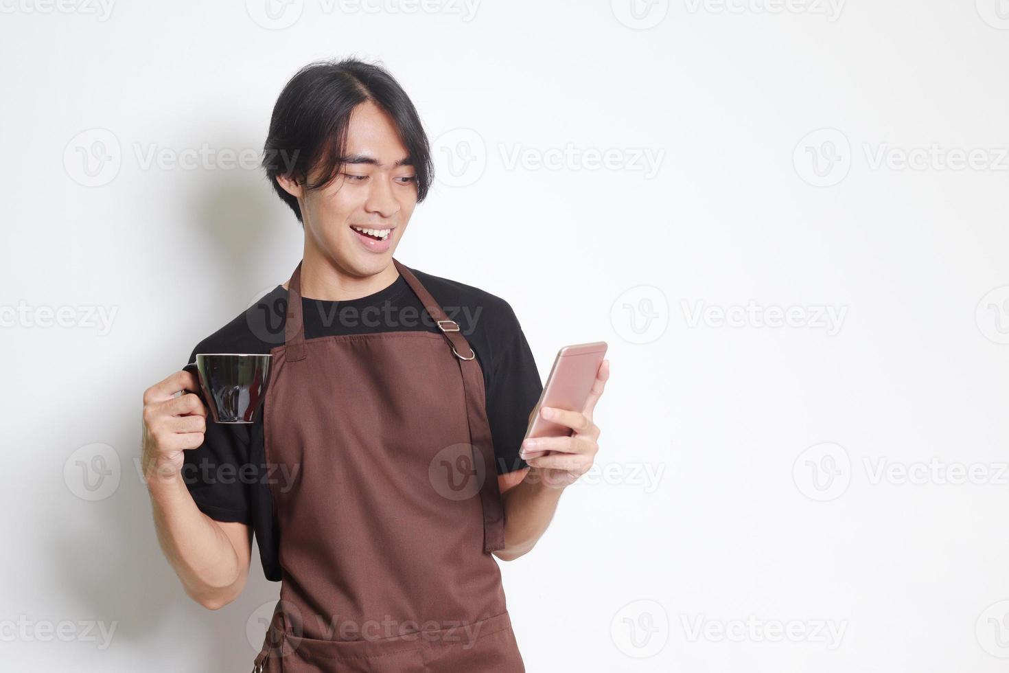 Portrait of attractive Asian barista man in brown apron holding a cup of coffee while using mobile phone and smiling at camera. Isolated image on white background photo