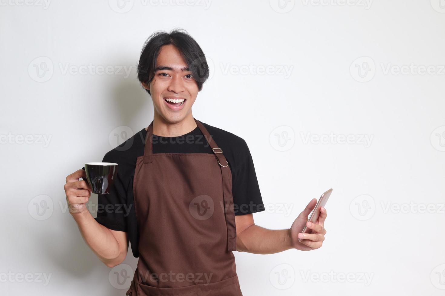 Portrait of attractive Asian barista man in brown apron holding a cup of coffee while using mobile phone and smiling at camera. Isolated image on white background photo