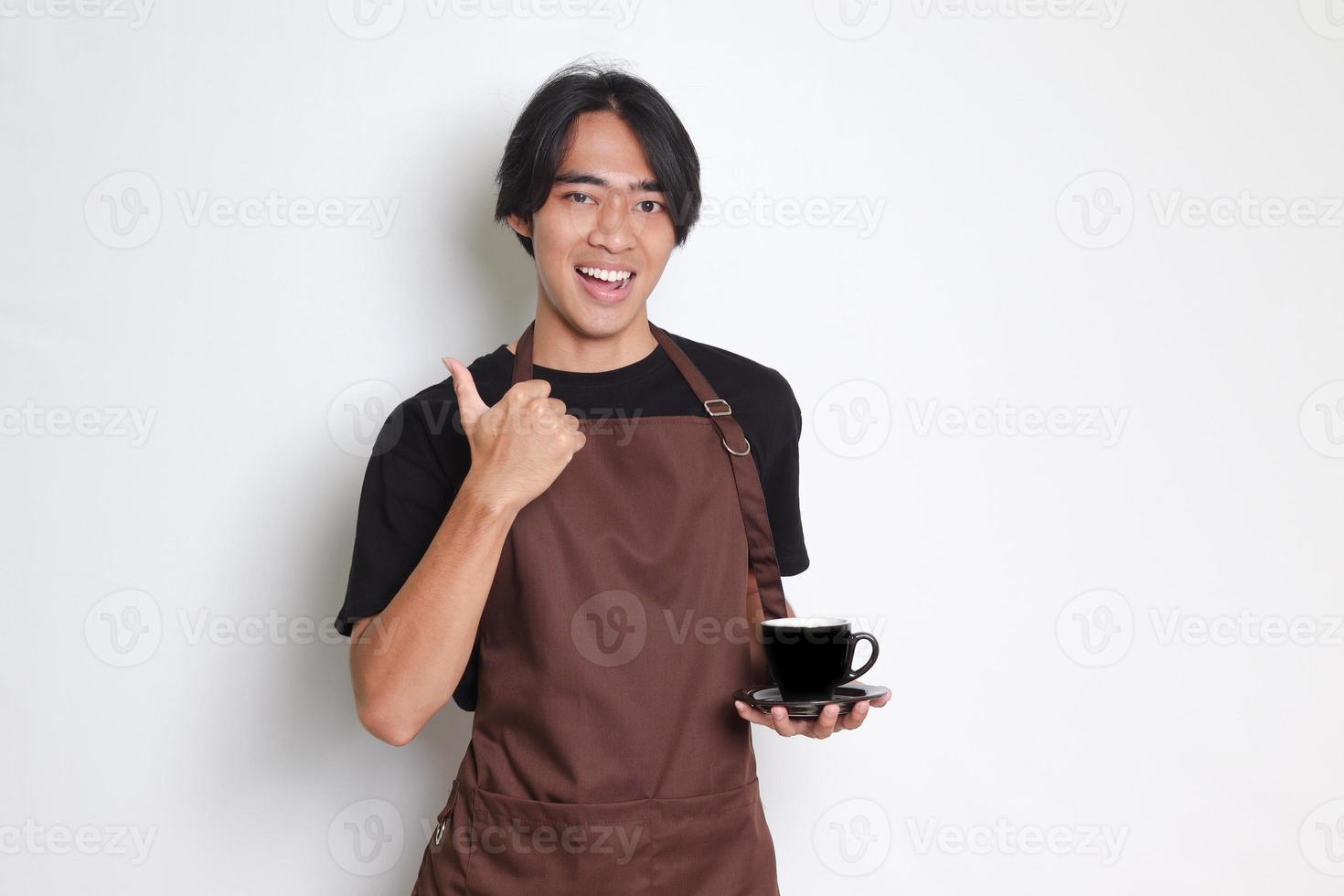 retrato de atractivo asiático barista hombre en marrón delantal servicio un taza de café a el cliente y haciendo pulgar arriba mano gesto. aislado imagen en blanco antecedentes foto