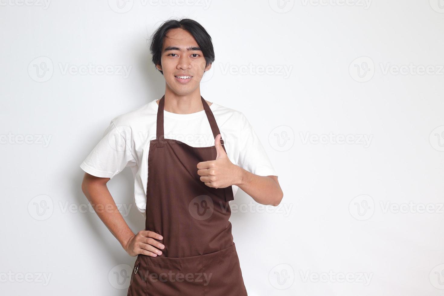 Portrait of attractive Asian barista man in brown apron showing thumb up hand gesture. Advertising concept. Isolated image on white background photo