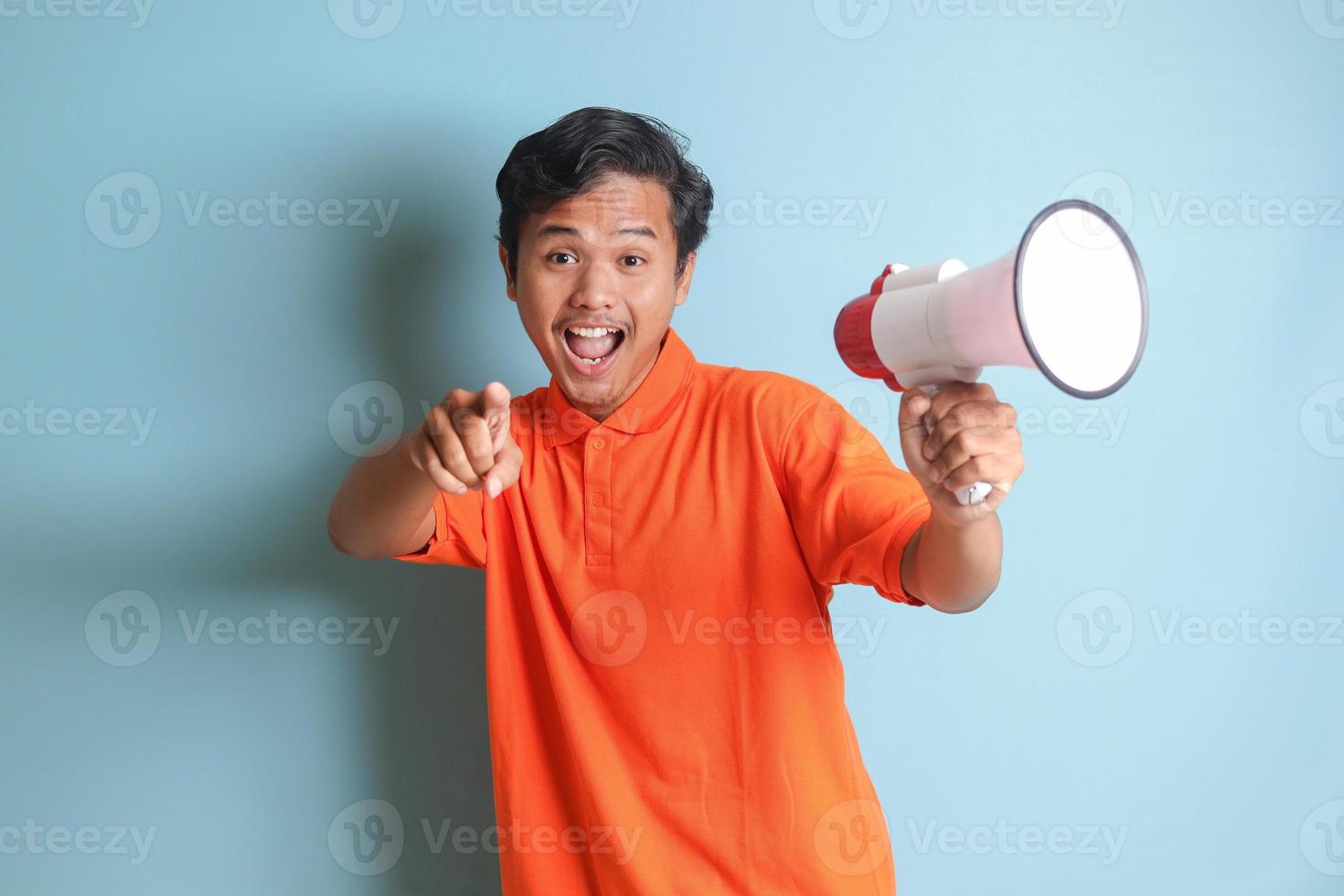 Portrait of attractive Asian man in orange shirt speaking louder using megaphone, promoting product. Advertising concept. Isolated image on blue background photo