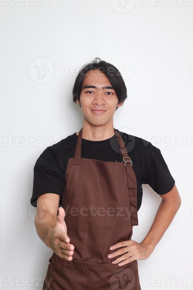retrato de atractivo asiático barista hombre en marrón delantal alcanzando fuera para un apretón de manos y saludar alguien. aislado imagen en blanco antecedentes foto