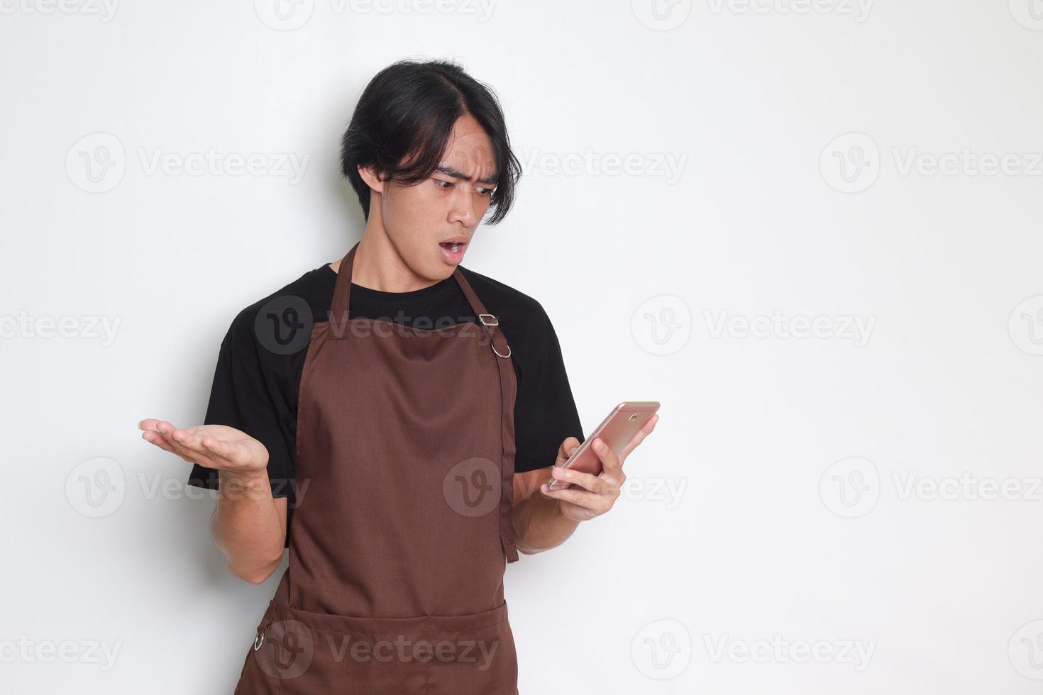 Portrait of shocked Asian barista man in brown apron looking at his mobile phone with surprised expression. Advertising concept. Isolated image on white background photo