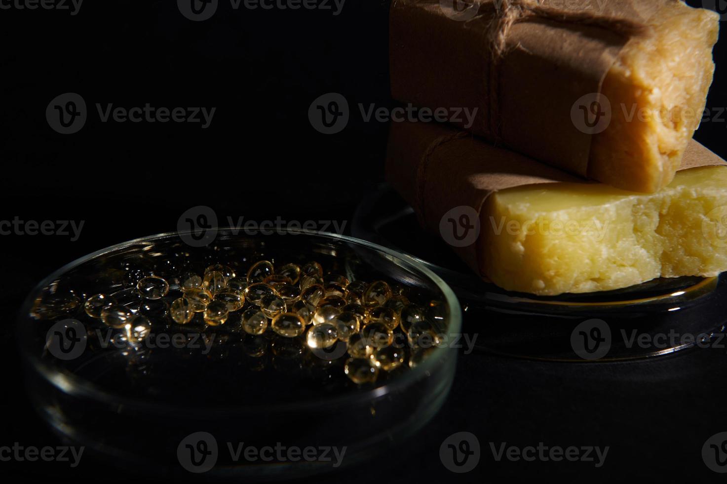 Translucent gel capsules with essential acids and oils in laboratory Petri dish against organic bars of soap on black photo