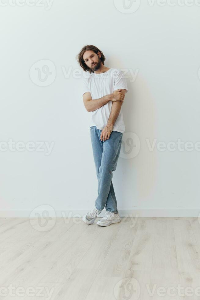 A man in a white T-shirt and blue jeans stands against a white wall and poses photo