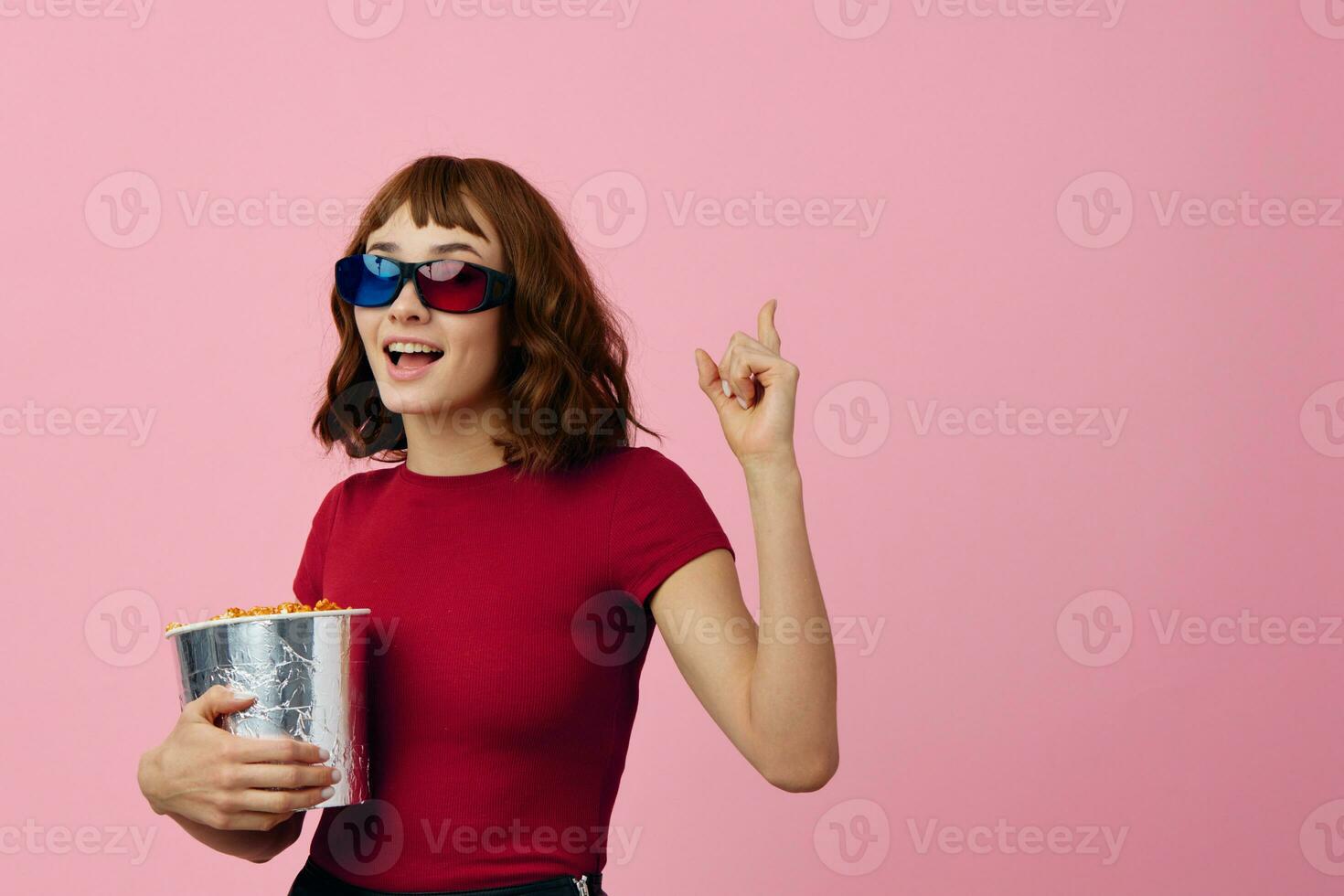 Overjoyed happy cute redhead lady in red t-shirt 3D glasses with popcorn watch movie posing isolated on over pink studio background. Copy space Banner. Fashion Cinema concept. Entertainment offer photo