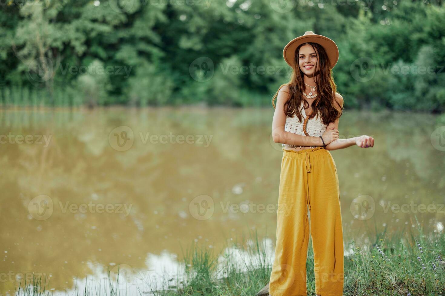 A young woman in a hippie look travels in nature by the lake wearing a hat and yellow pants in the fall photo