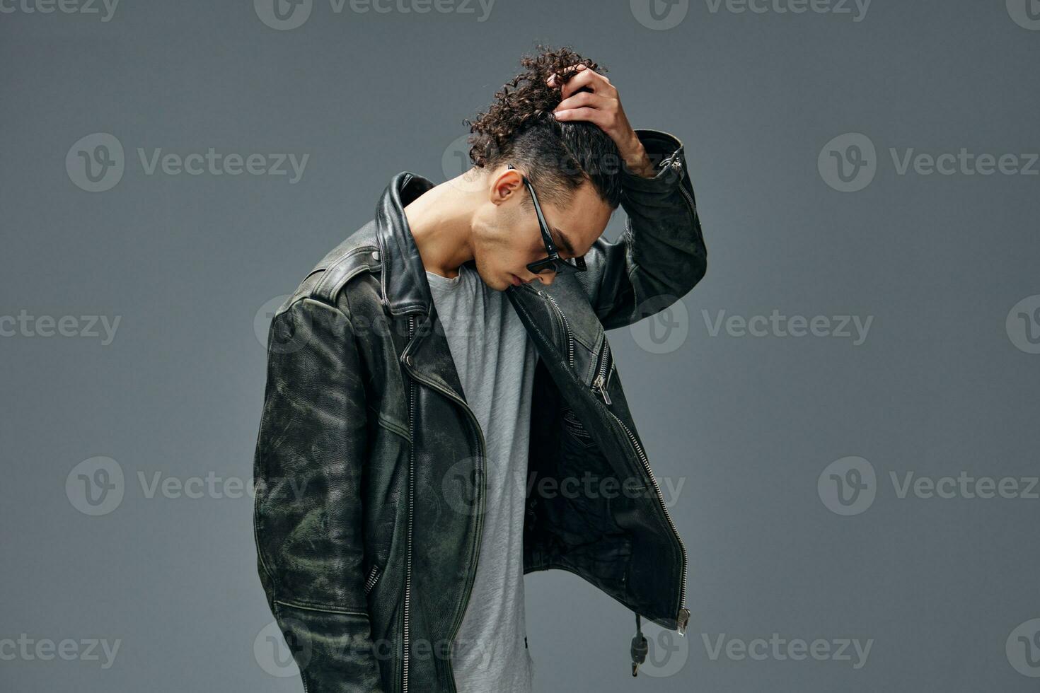 Side of view of stylish tanned curly man leather jacket trendy sunglasses posing isolated on gray studio background. Cool fashion offer. Huge Seasonal Sale New Collection concept. Copy space for ad photo