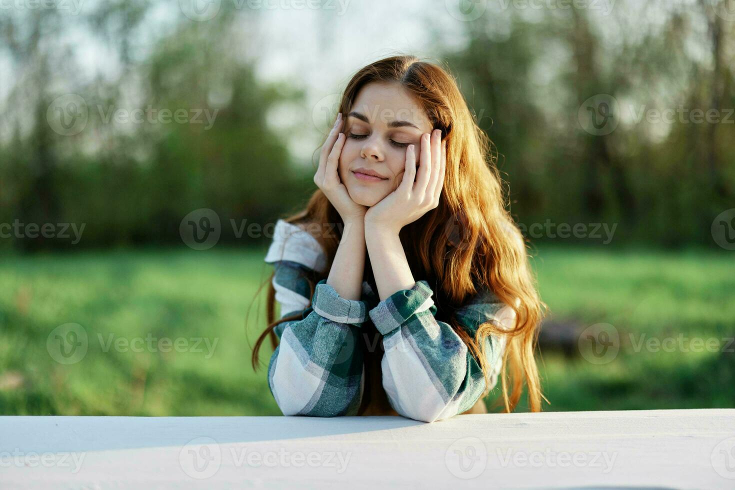 A beautiful young woman with long curly red hair in a plaid shirt sits with her hands under her head in the summer greenery photo