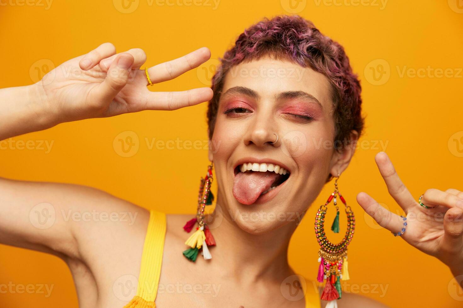 Portrait of a young woman with a short haircut and colored hair smiling and showing her tongue at the camera on an orange background with earrings accessories in the studio photo