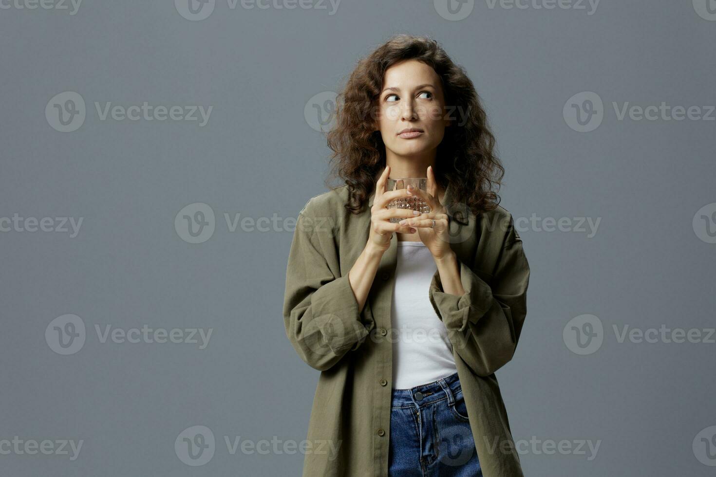 Dreaming curly beautiful woman in casual khaki green shirt hold glass of water thinking looks aside posing isolated on over gray blue background. Healthy lifestyle. Water is Life concept. Copy space photo