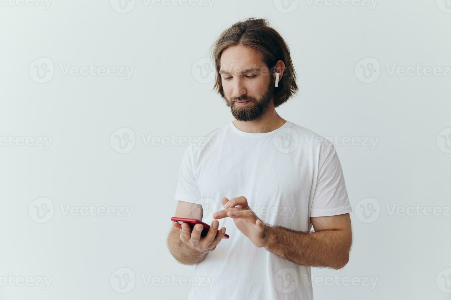 A man with a beard and long hair in a white T-shirt and blue jeans looks at his phone flipping through an online social media feed with headphones in his ears against a white background photo