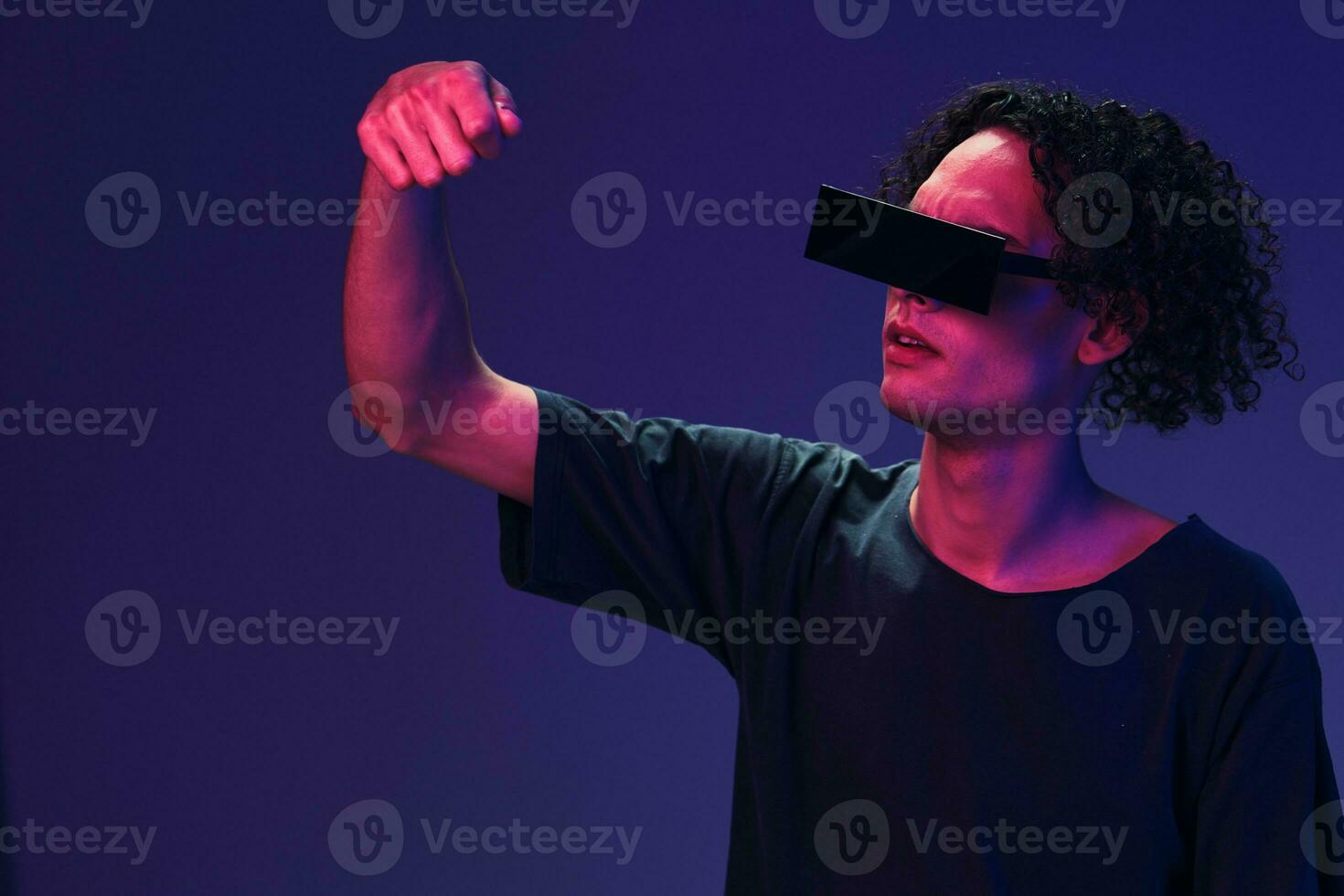 Excited stylish tanned curly man in black t-shirt eyewear holds invisible object posing isolated on color pink blue background. Cool fashion offer. Virtual Reality New Collection concept. Copy space photo