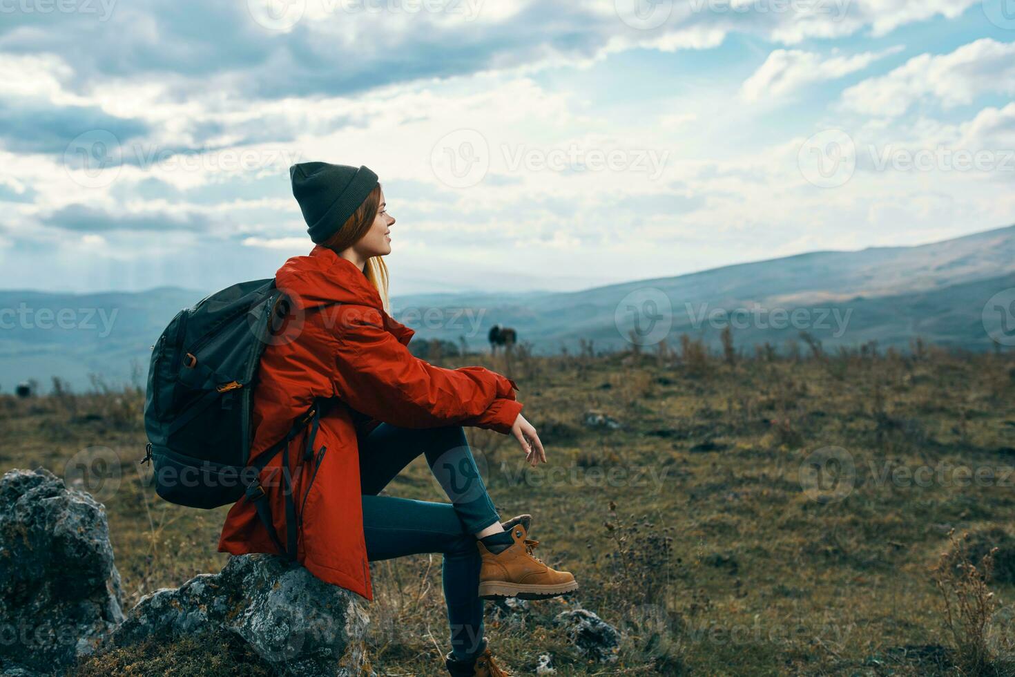 Traveler In a red jacket, hats with a backpack sits on a stone in the mountains in nature photo