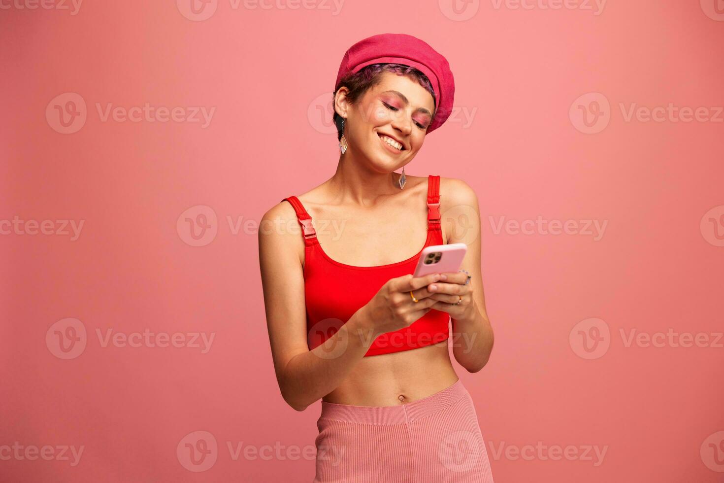 Young woman blogger with colored pink hair and short haircut flipping through the phone screen and typing a message with a smile and surprise in stylish clothes on a pink background photo