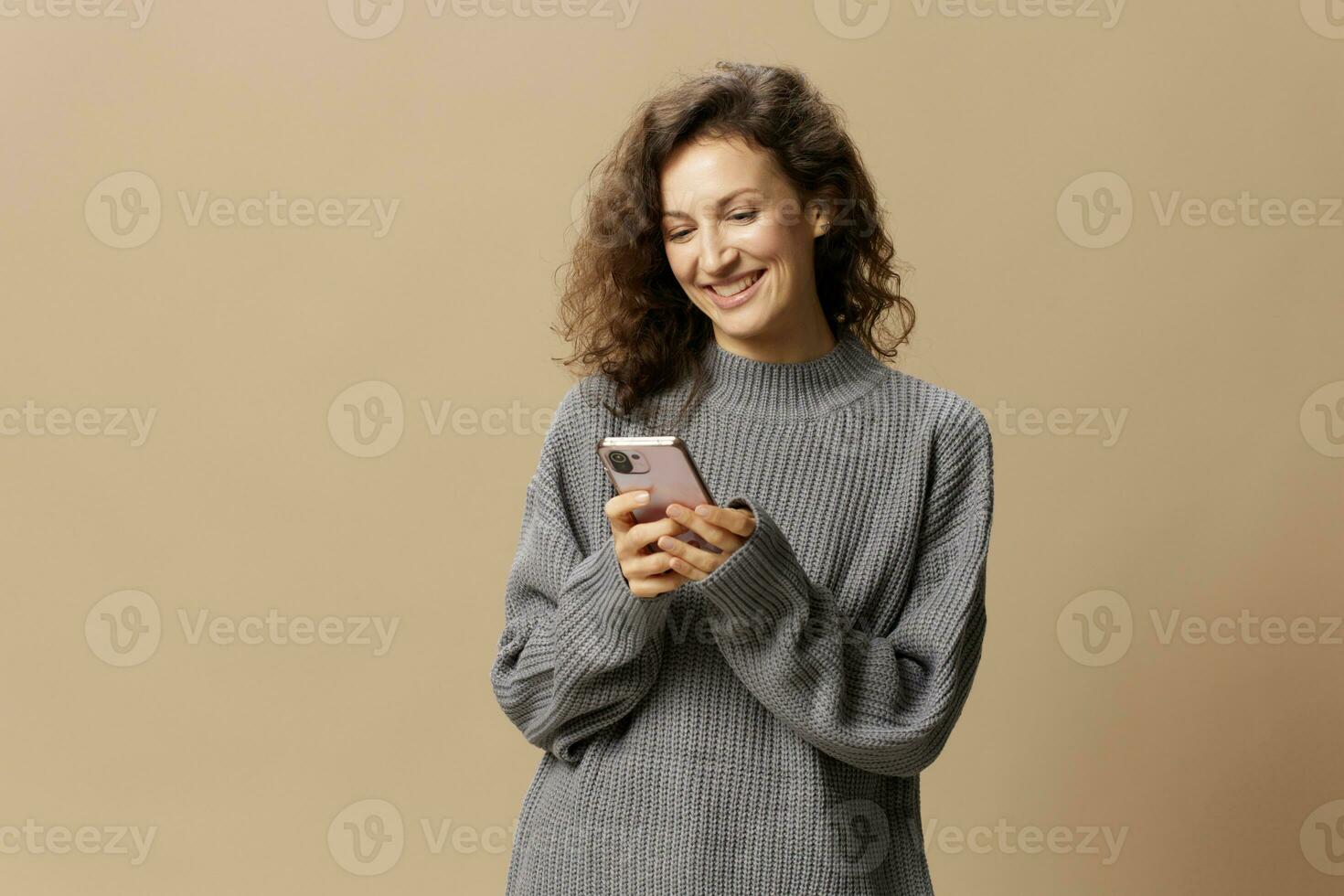 Lovely happy curly beautiful female in gray casual sweater chatting with boyfriend posing isolated on over beige pastel background. Social media, network, distance communication concept. Copy space photo