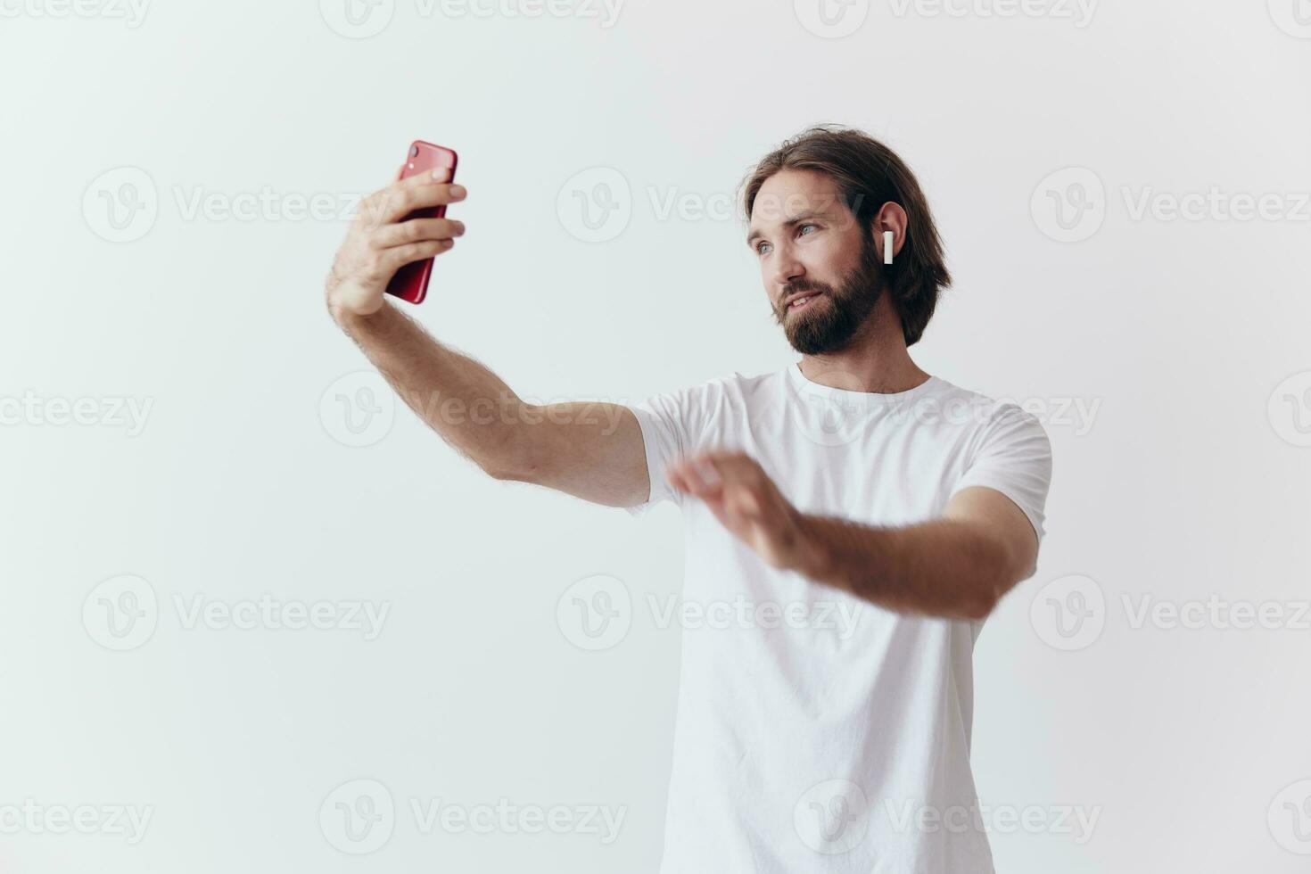 A male blogger videotaping himself on his phone and chatting with people online with a smile in a white T-shirt against a white wall photo